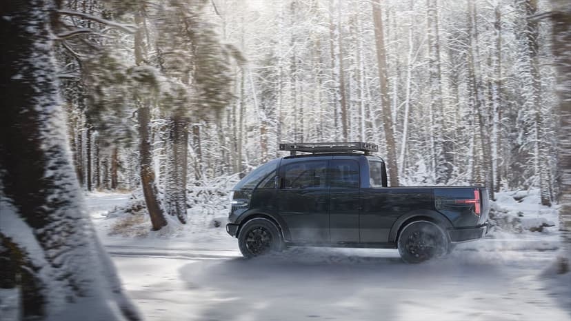 Black electric pickup truck driving on a snowy woodland road surrounded by frost-covered evergreen trees.