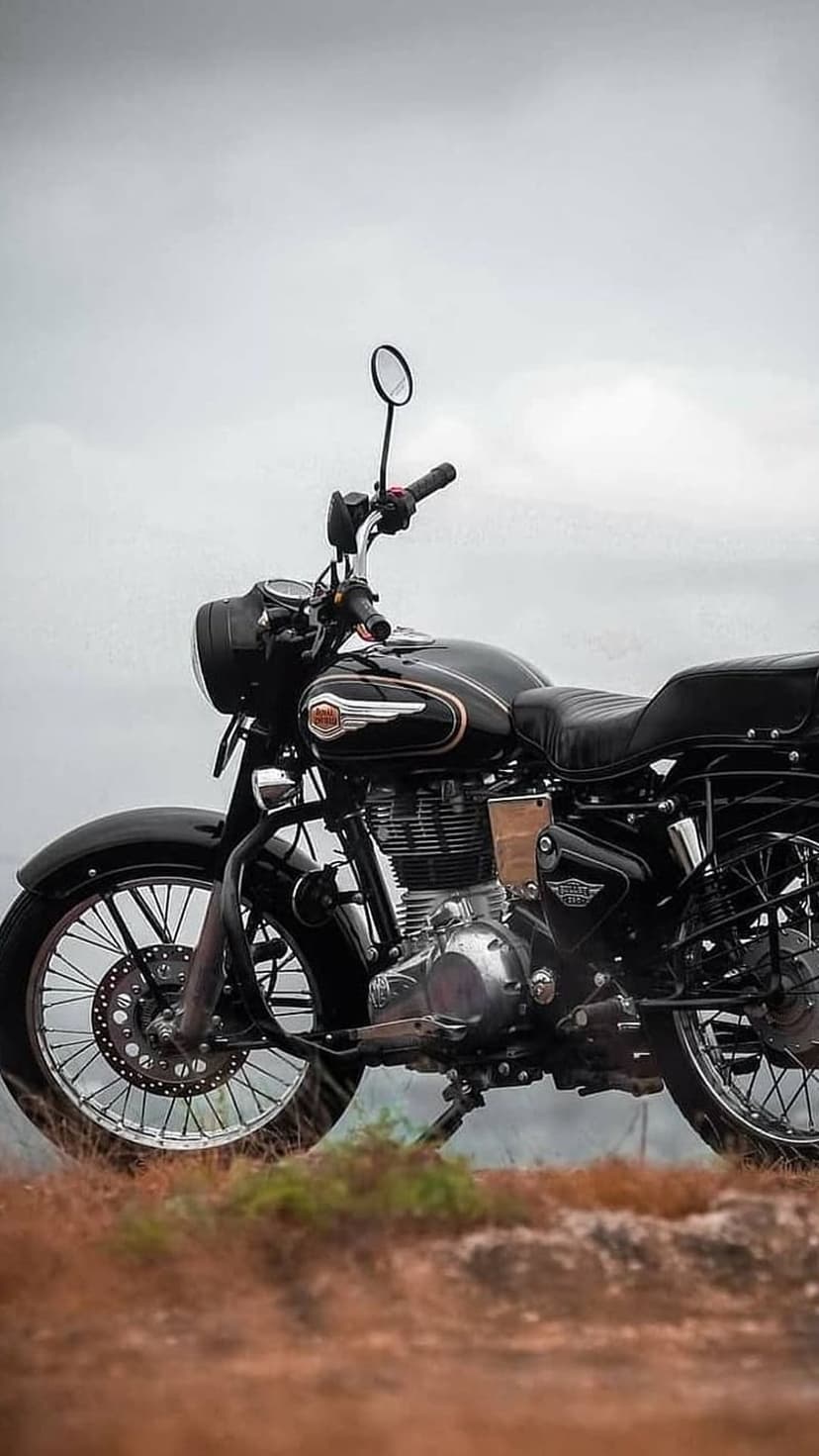 Black Royal Enfield motorcycle with chrome engine on a grassy hill under a cloudy sky in a vertical shot.