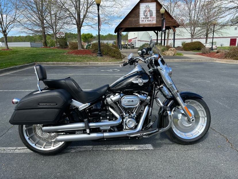 Black Harley-Davidson Fat Boy 114 cruiser with chrome exhaust parked in a lot under a clear sky.