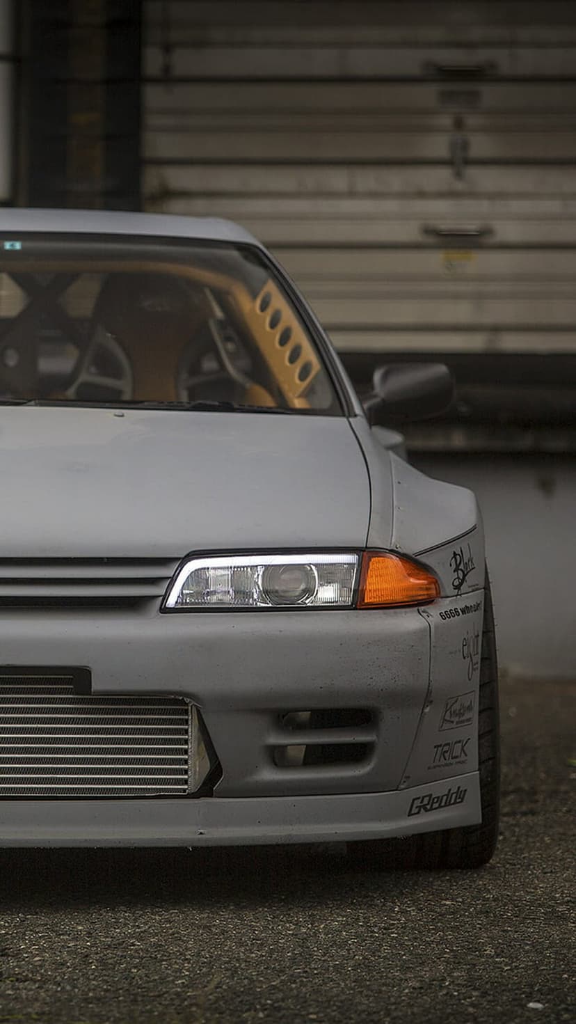 Nissan Skyline R32 GT-R with a matte grey body, exposed intercooler, and LED headlights in front of a metal door.