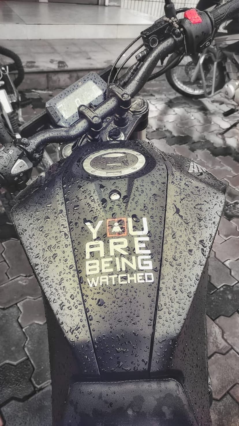 Black motorcycle fuel tank covered in raindrops with a white You Are Being Watched decal and orange icon.