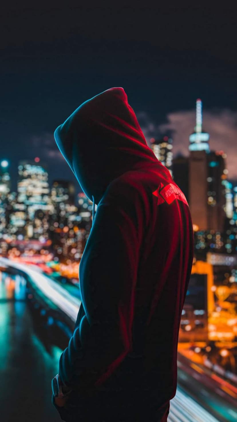 Person in a dark hoodie stands before red and blue neon light trails on a blurred urban bridge at night.