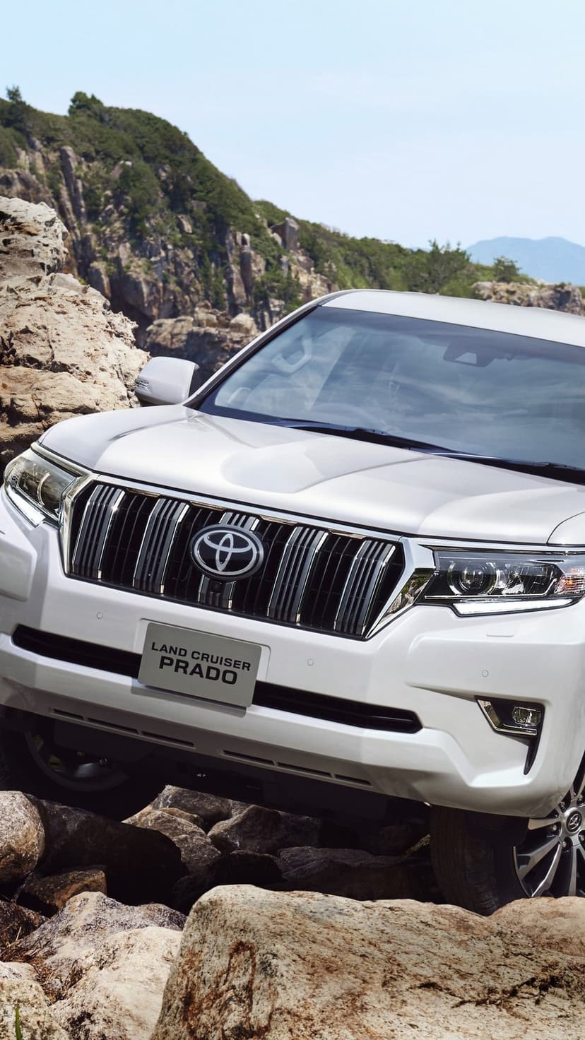 White Toyota Land Cruiser Prado SUV parked on a rocky cliffside with mountains and blue sky background.