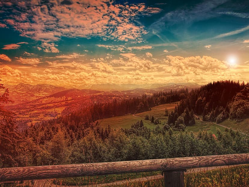 Rolling mountains under an orange sunset sky with a rustic wooden fence framing the foreground landscape.