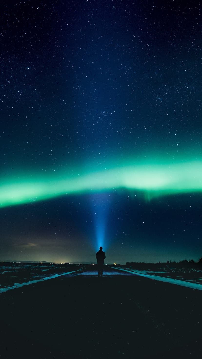 Lone person on a dark road shines a vertical light beam toward swirling green northern lights and a starry sky.