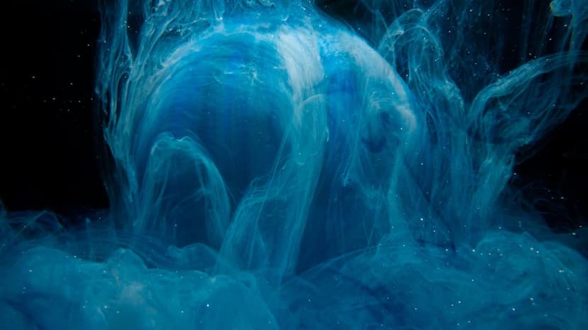 Glowing white sphere surrounded by turbulent swirls of cobalt blue ink and fine particles on a dark backdrop.