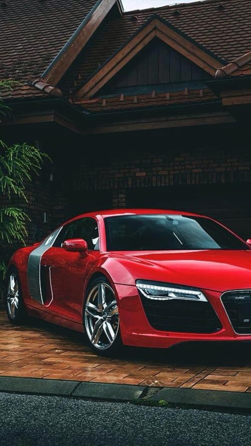 Red Audi R8 sports car parked on a brick paver driveway in front of a modern house with lush green plants.