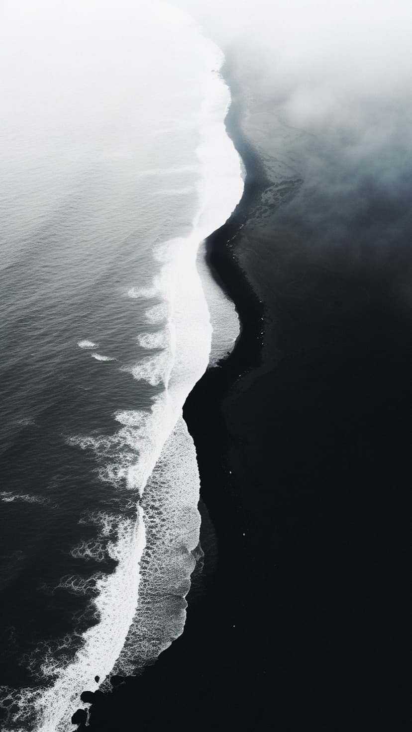 Black sand beach meets white frothy ocean waves in an aerial view showing sharp contrast and misty textures.