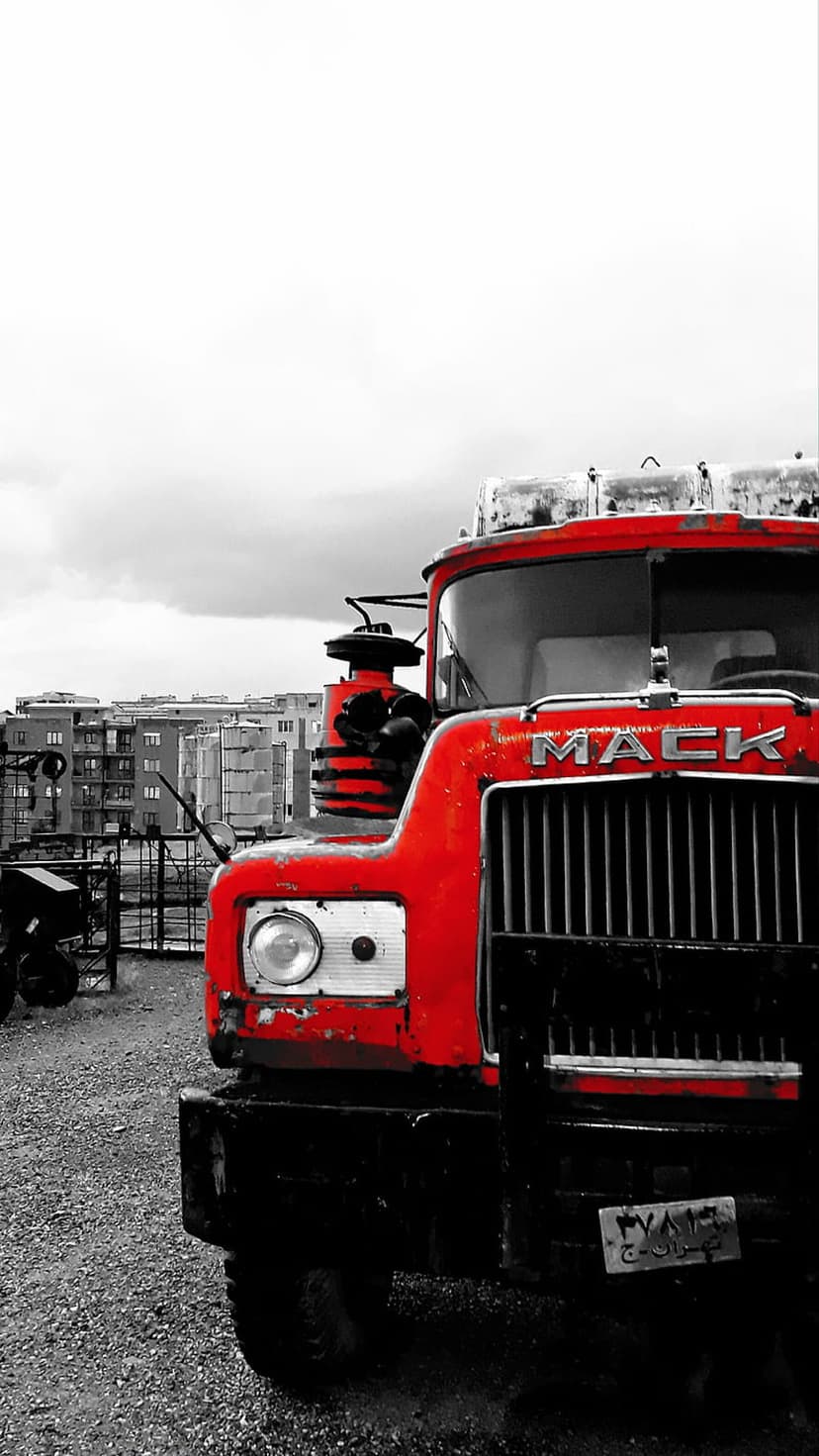 Red Mack truck with a chrome grille and weathered paint parked in a grayscale urban industrial environment.