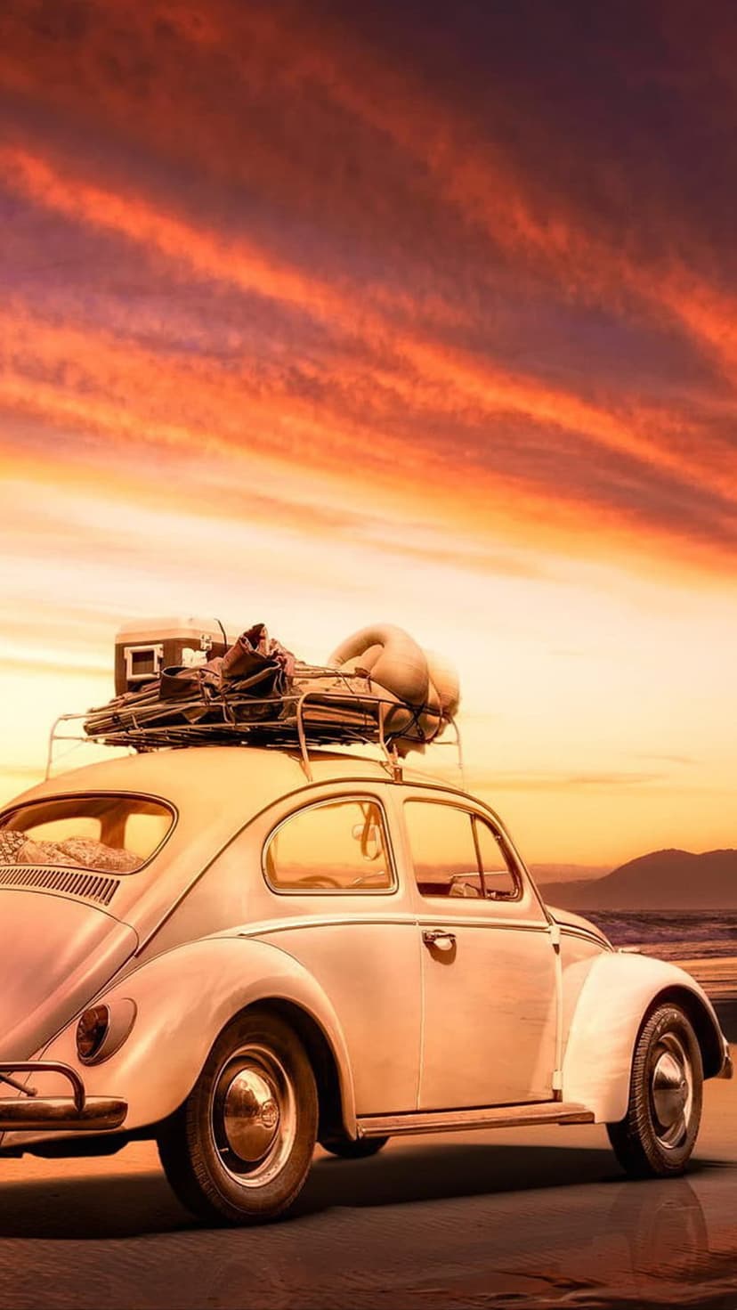 White Volkswagen Beetle with luggage on its roof rack driving on a sandy road during a vibrant beach sunset.