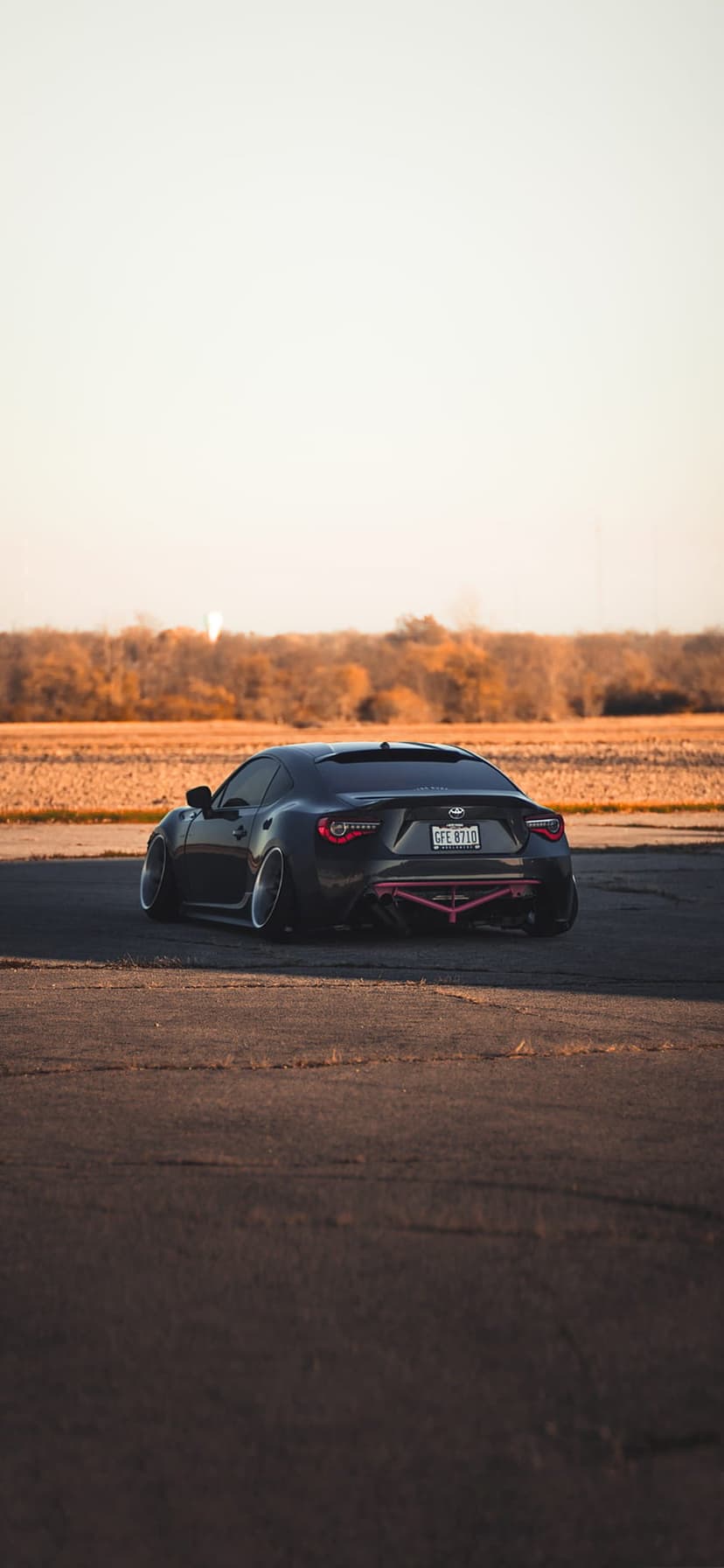 Black Subaru BRZ sports car with a pink rear diffuser on asphalt amidst golden autumn foliage during sunset.