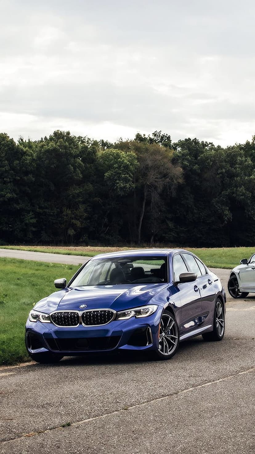 Blue BMW sedan parked on a paved road beside a green forest under a cloudy sky with a car in the background.