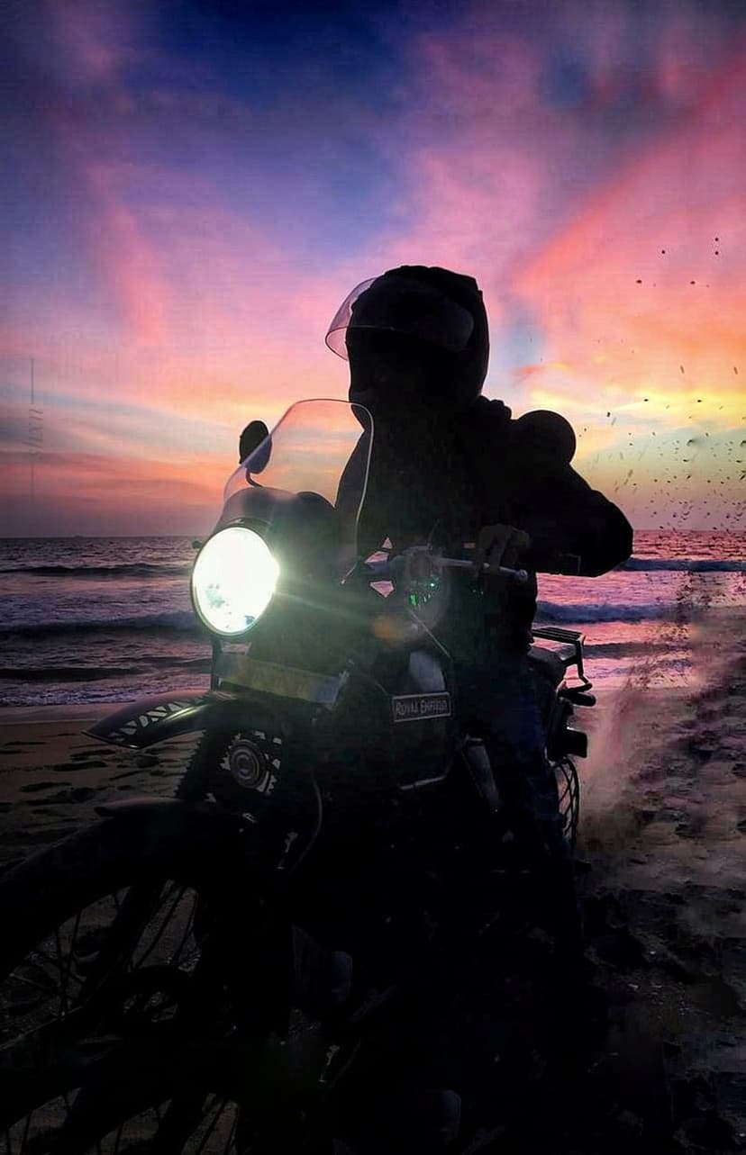 Motorcyclist silhouette on a wet beach during a pink and orange sunset with waves crashing in the background.
