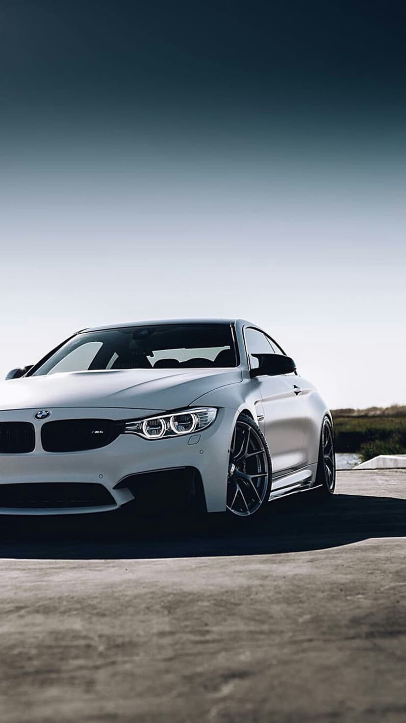 White BMW M4 Coupe with black wheels parked on an asphalt road beneath a clear blue sky and distant trees.