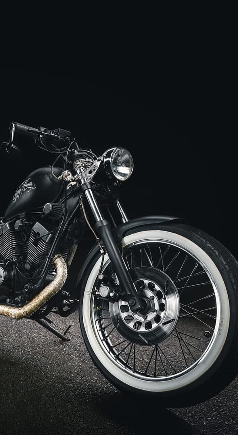 Custom black motorcycle featuring a chrome headlight and whitewall tires parked on asphalt against a dark background.