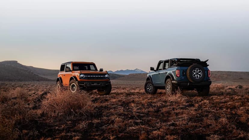 Orange and blue Ford Bronco SUVs parked on rocky desert terrain under a soft sky with distant mountains.