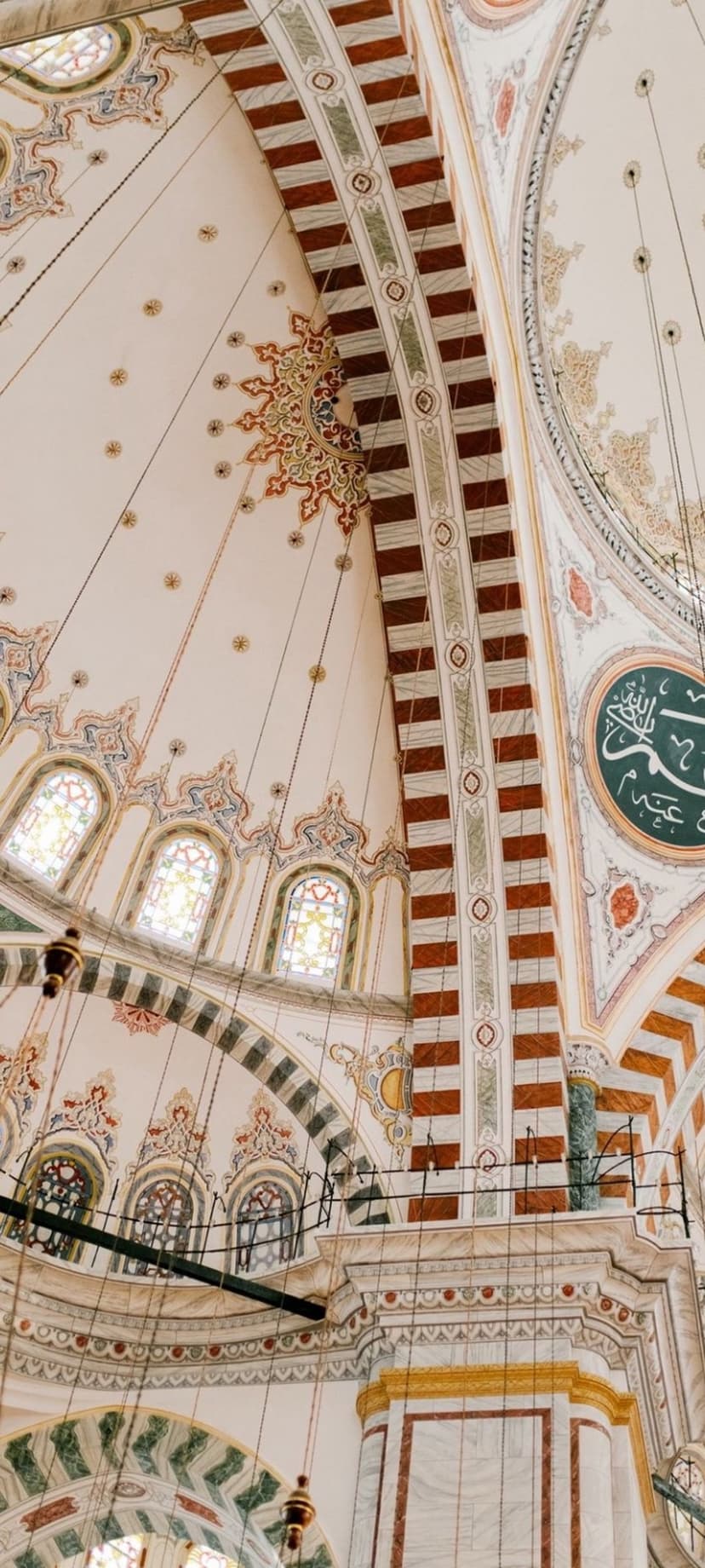 Islamic dome ceiling with gold calligraphy, geometric patterns, and arched stained glass windows in a mosque.
