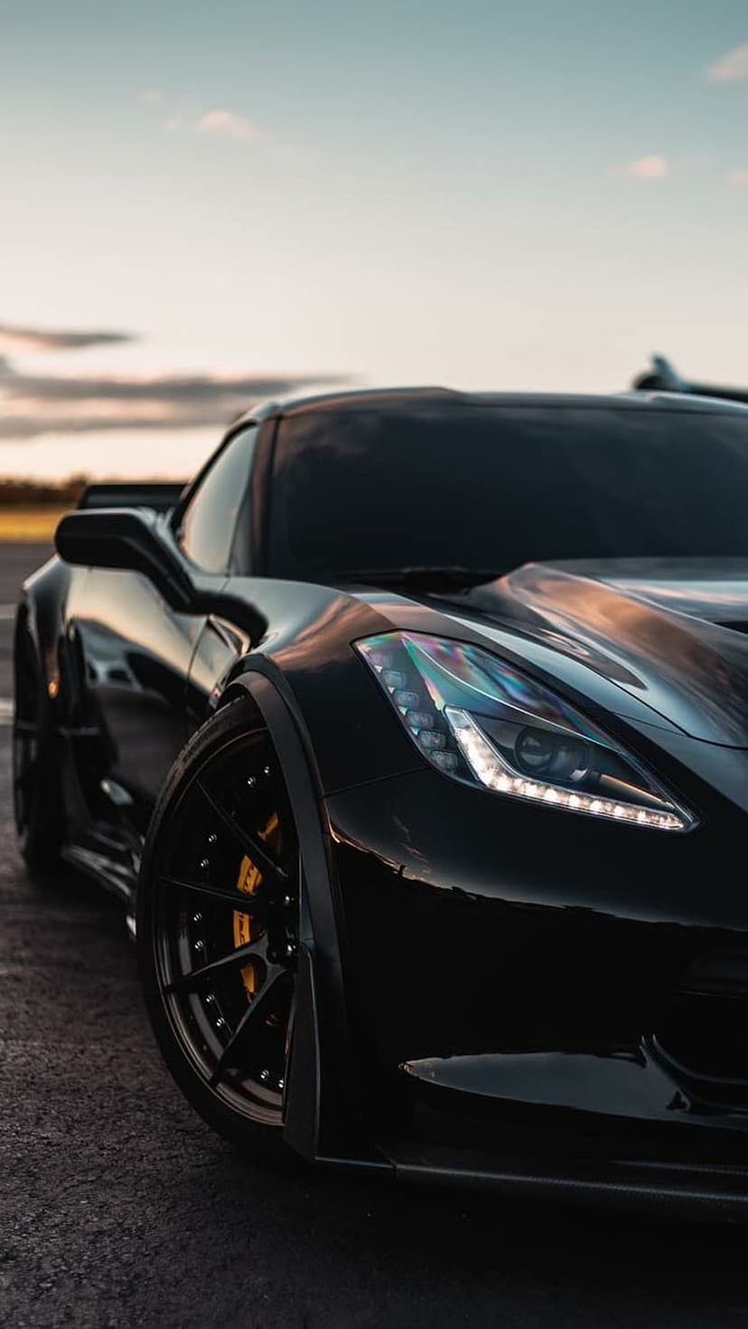 Black Corvette C7 with yellow brake calipers parked at sunset in a low-angle vertical automotive photograph.