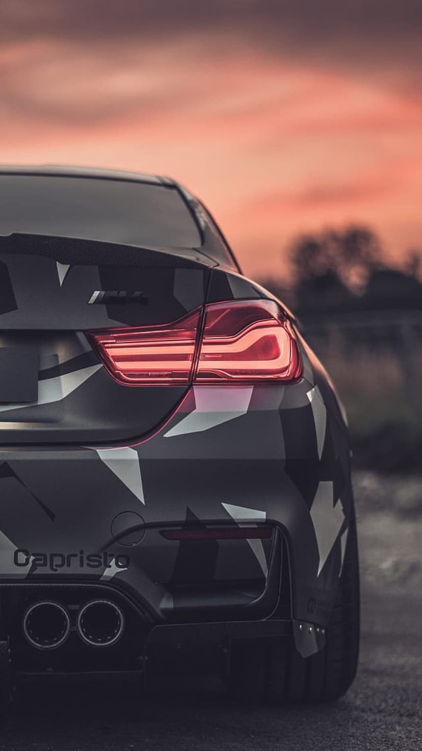 BMW M4 rear view featuring a camouflage wrap, glowing red taillights, and dual exhausts against a sunset sky.