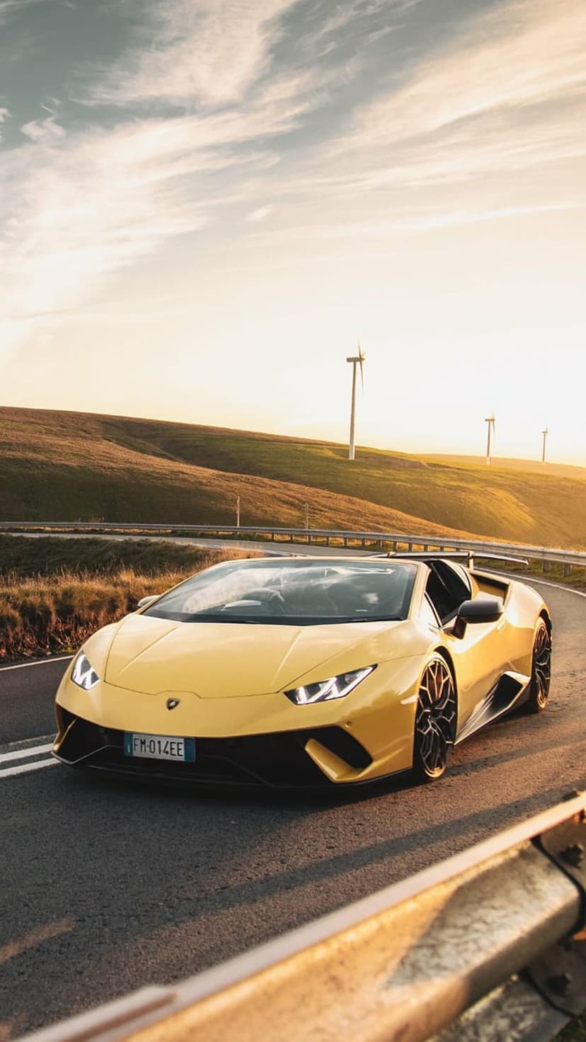 Yellow convertible Lamborghini driving on a winding road past wind turbines under a warm golden sunset sky.