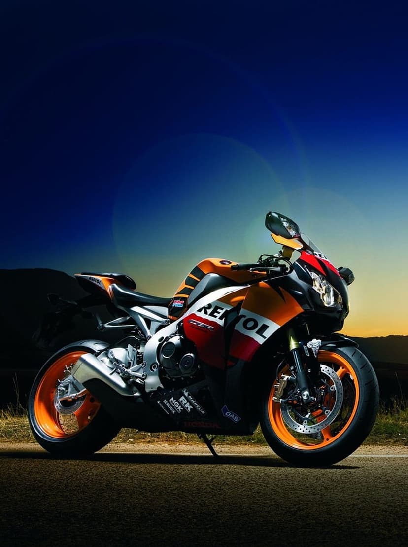 Repsol Honda CBR motorcycle on a mountain road at sunset with orange racing fairings and a golden sky background.