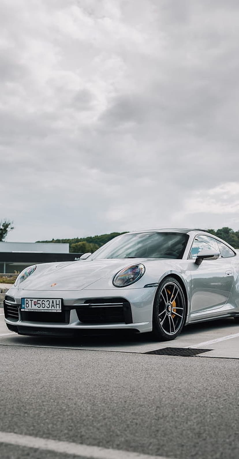 Silver Porsche 911 Turbo S with yellow brake calipers parked on pavement under a moody cloudy sky.