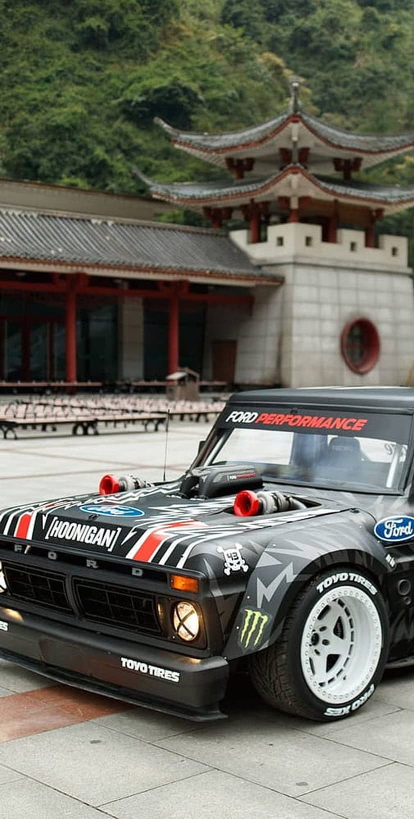 Modified Ford truck with twin turbos parked near a red Asian pagoda with a lush green mountain in the background.