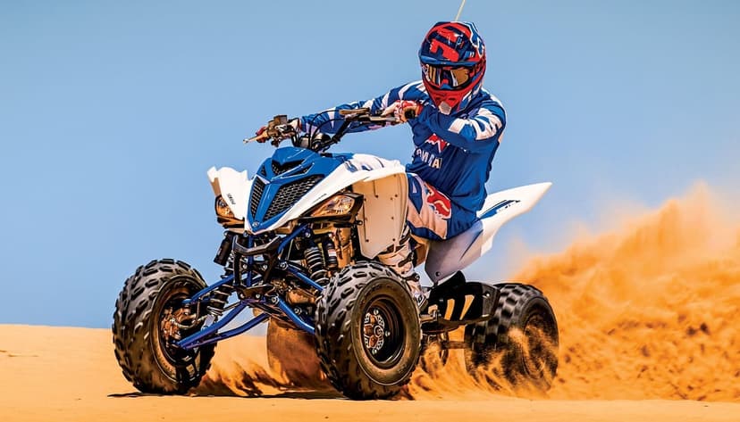 Yamaha Raptor ATV racing across desert dunes, kicking up sand with a rider in blue gear under a bright blue sky.