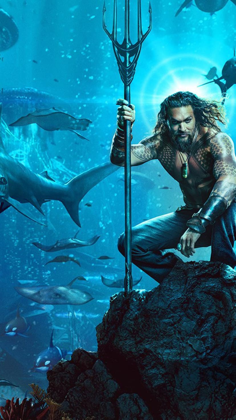 Aquaman holds a golden trident while standing on a reef surrounded by swimming sharks and rays in blue ocean water.