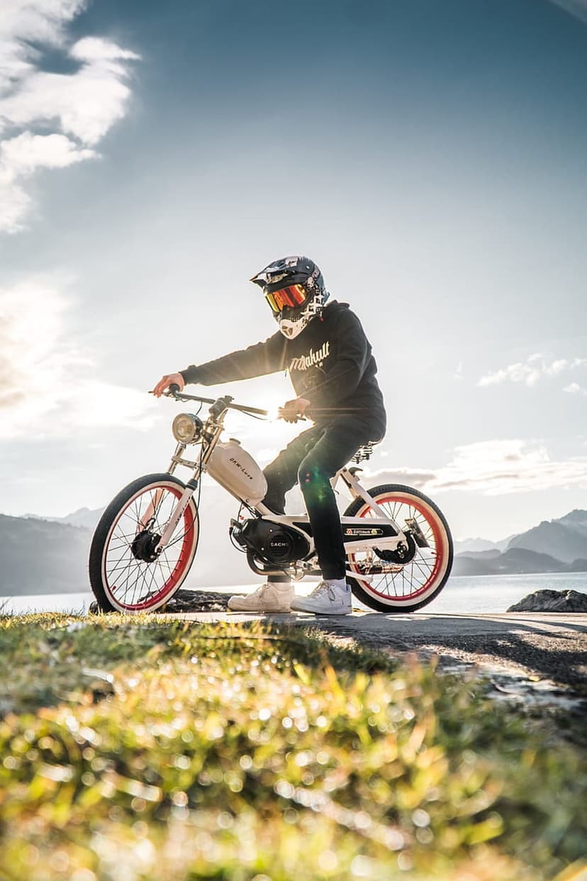 Moped rider in a helmet sits by a scenic mountain lake with a bright golden sun flare on a grassy roadside.