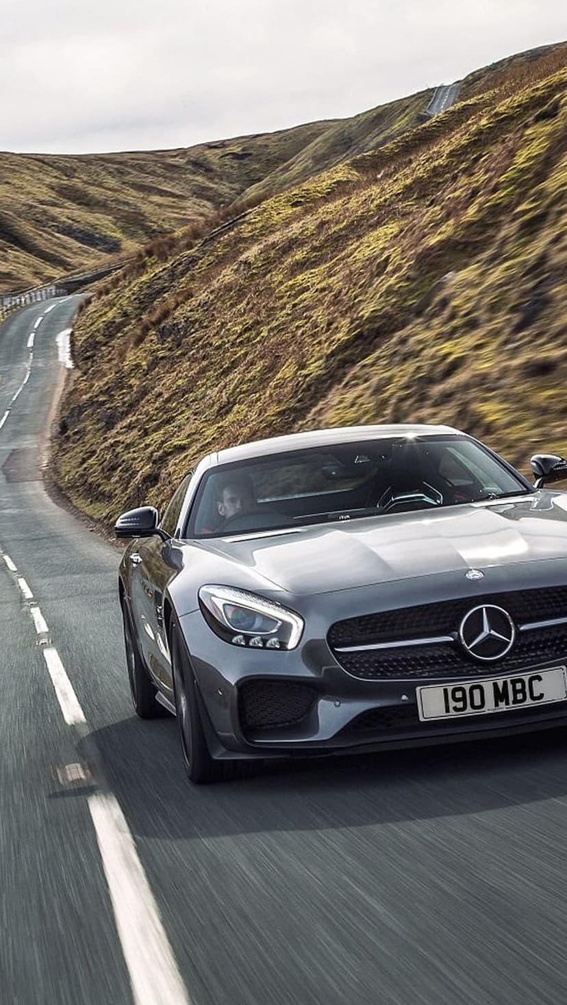 Mercedes-AMG GT S drives on a winding mountain road under an overcast sky surrounded by rolling hills.