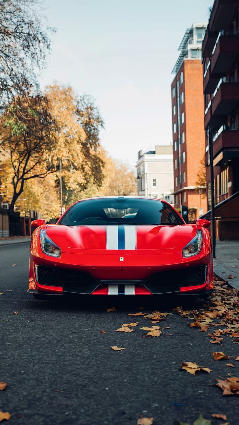 Red Ferrari with racing stripes parked on a city street covered in golden autumn leaves near modern buildings.