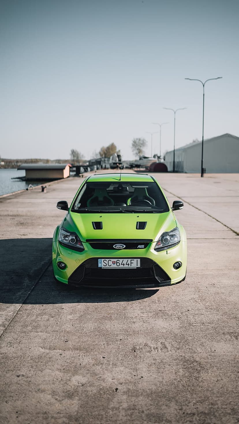 Lime green Ford Focus RS parked on a concrete pier next to calm water with industrial buildings and a blue sky.