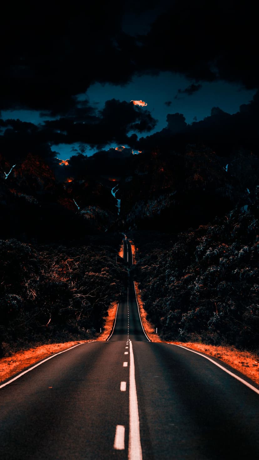 Vanishing asphalt road flanked by dark tree silhouettes against a dramatic blue and orange storm sky.