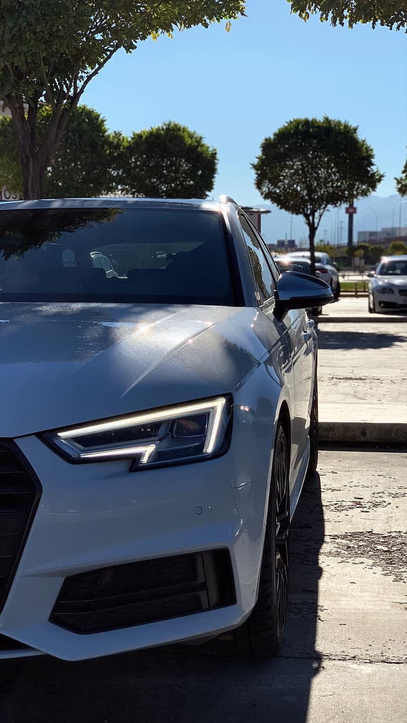 White Audi wagon with glowing LED headlights parked in a sunny lot under a blue sky and green trees.