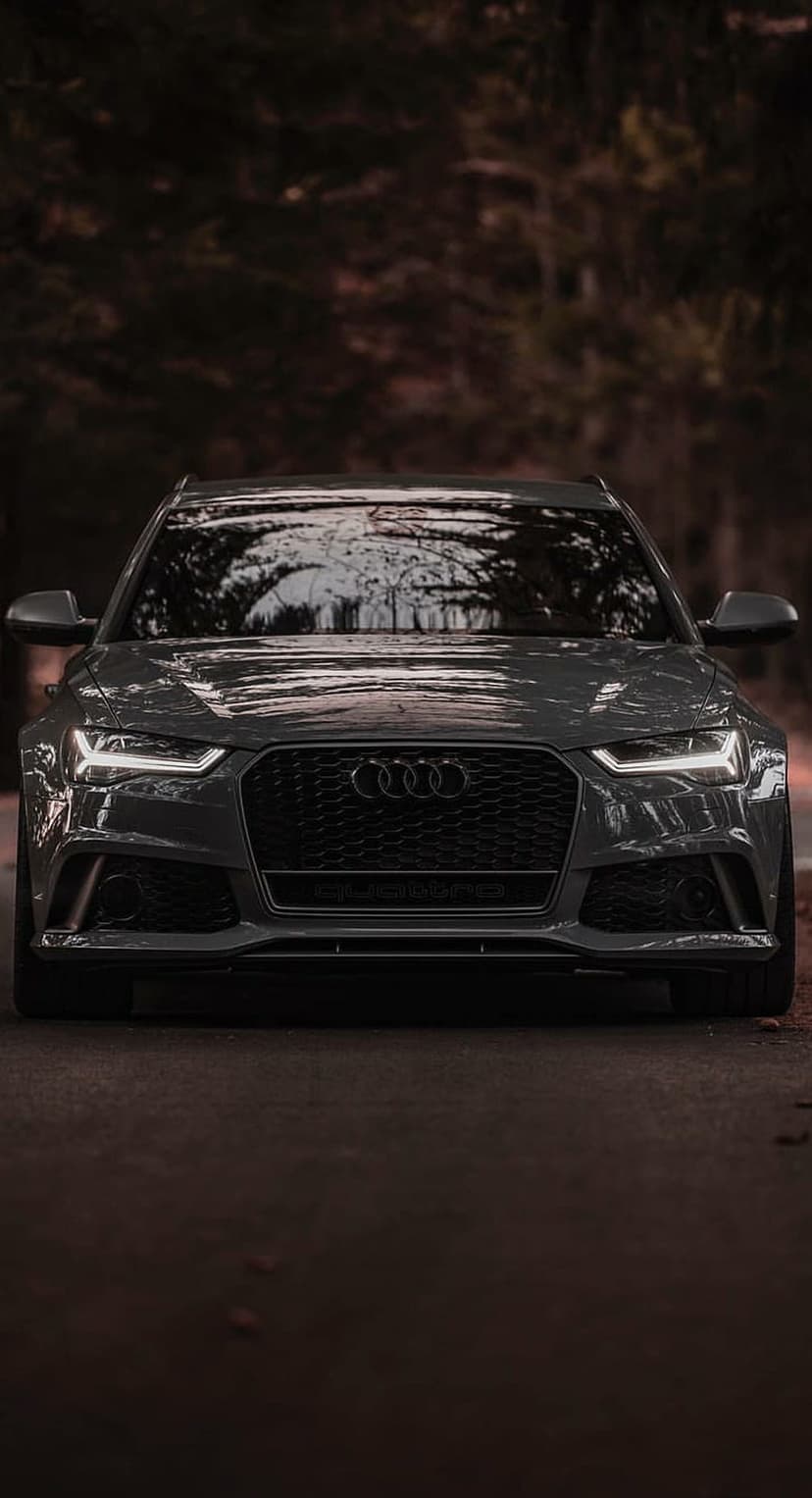 Audi RS6 Quattro front view with glowing headlights on a forest road under dim, atmospheric lighting.