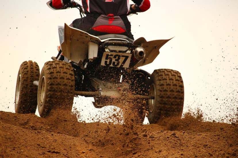 Quad bike rider wearing protective gear races through loose dirt, throwing a spray of earth behind the vehicle.