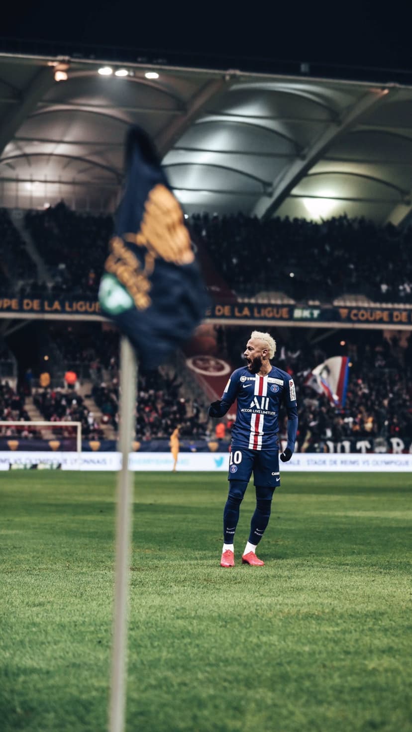 Neymar Jr. stands on a grass field in a PSG jersey under bright stadium lights with a blurred flag in front.