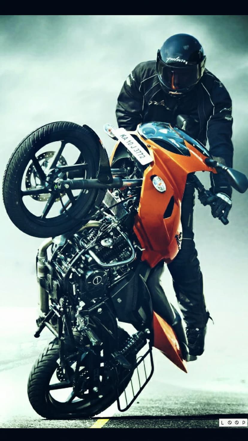 Motorcyclist on an orange sport bike performs a wheelie against a hazy background, wearing black riding gear.