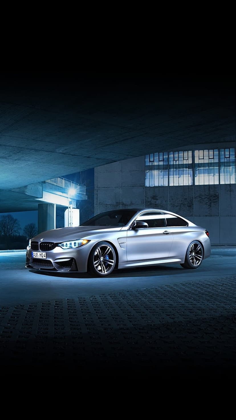 Silver BMW M4 coupe parked in a dark concrete warehouse with dramatic blue lighting and industrial windows.