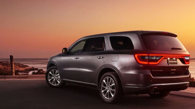 Gray Dodge Durango SUV with glowing red taillights parked on a coastal road overlooking a sunset ocean.