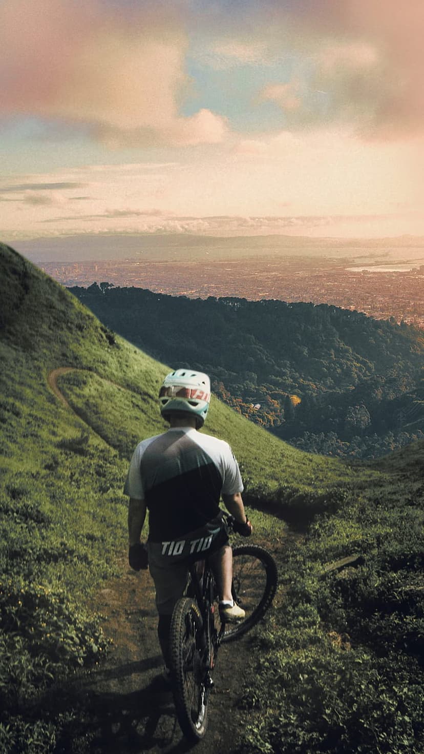 Mountain biker in a helmet stands on a grassy trail overlooking a vast city during a soft golden hour sunset.