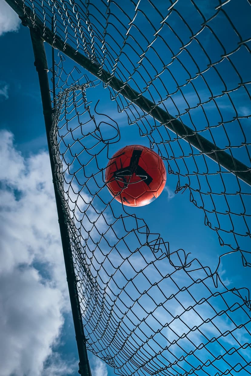 Orange soccer ball passes through a hole in a chain-link fence against a bright blue sky with white clouds.