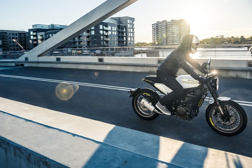 Motorcyclist on a white Husqvarna Vitpilen riding across a bridge at sunset with city buildings and lens flares.