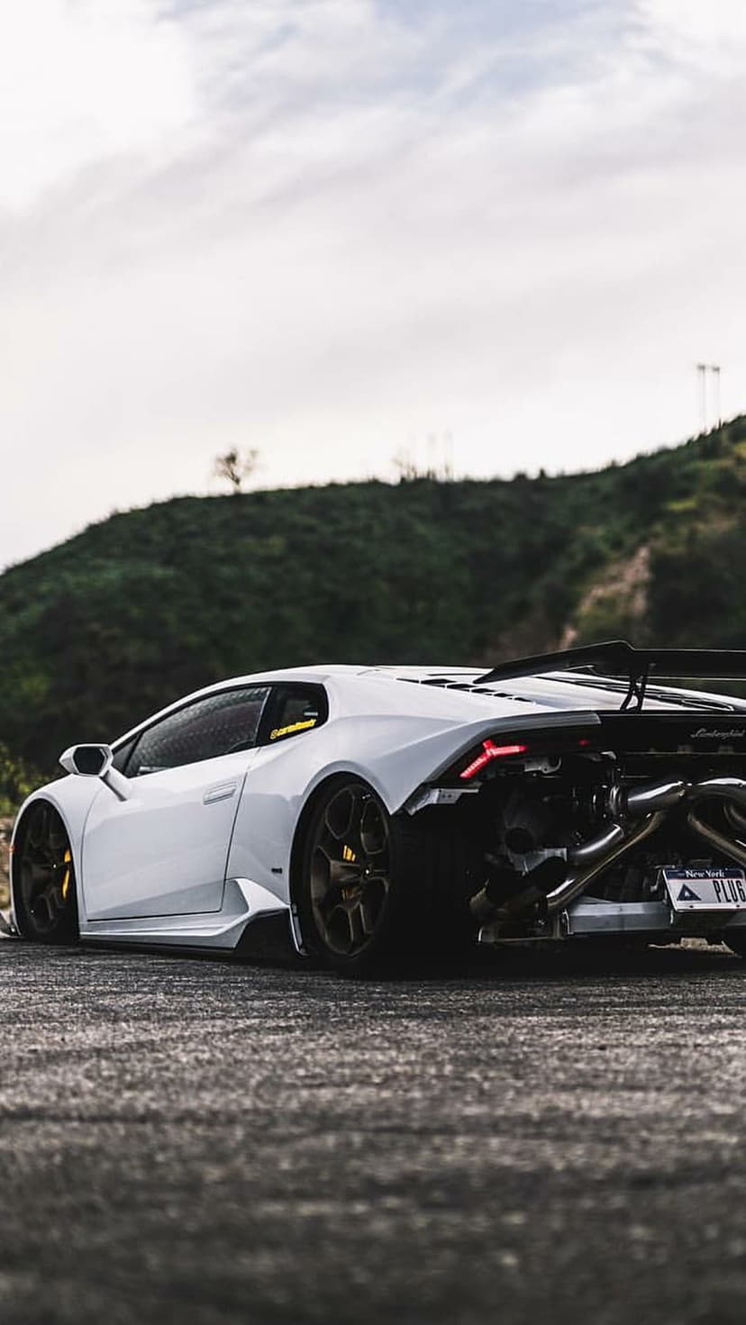 White Lamborghini Huracan with an exposed engine and custom exhaust parked on a road near rolling green hills.