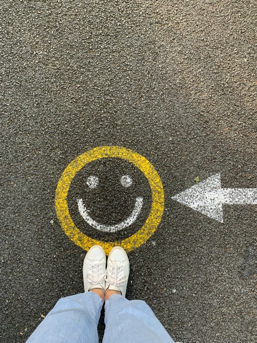 Yellow smiley face and white arrow painted on asphalt with white sneakers standing at the bottom edge.