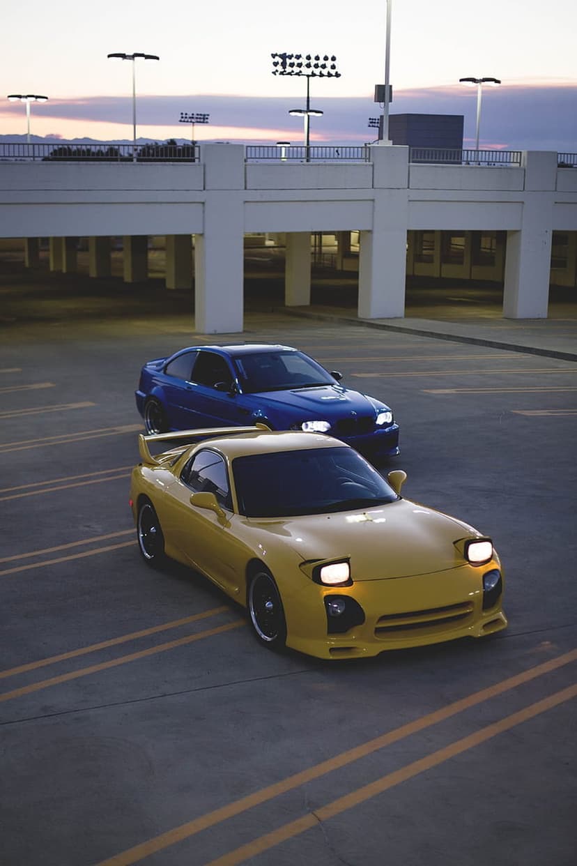 Yellow Mazda RX-7 with pop-up lights on parked beside a blue BMW in a concrete parking garage at twilight.