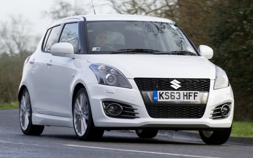 White Suzuki Swift Sport hatchback driving on an asphalt road with a blurred green forest background.