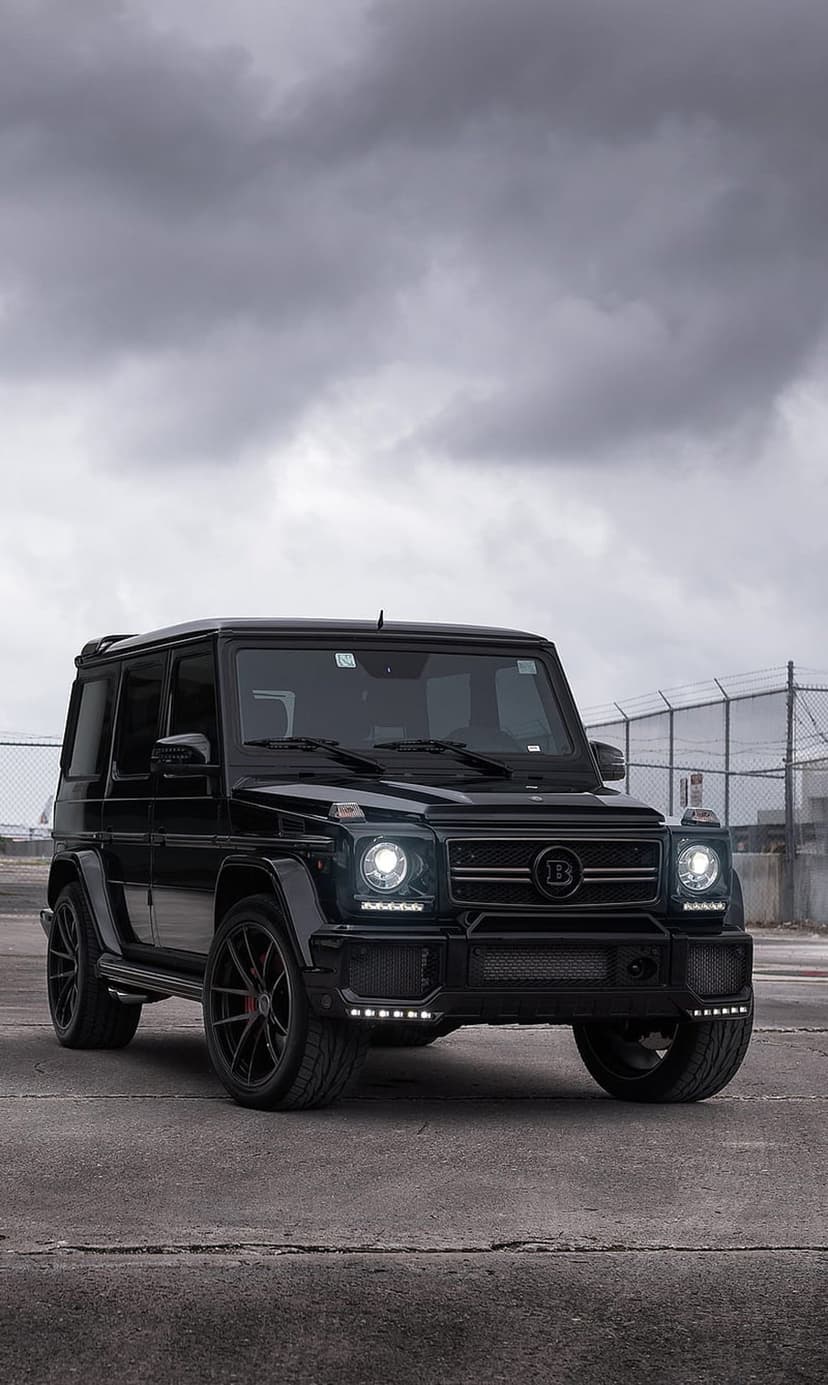 Black Brabus G-Wagon SUV with illuminated LED headlights parked under a heavy grey sky in a vertical shot.