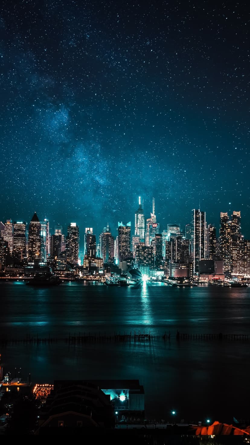Manhattan skyscrapers reflect in dark harbor water beneath a star-filled Milky Way galaxy in the night sky.